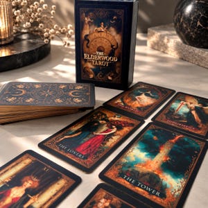 Könnte beinhalten: Ein Kartendeck The Elderwood Tarot mit einer dunklen Schachtel mit goldener Schrift und mehreren ausgebreiteten Karten, darunter The Lovers, The Tower, The World und Judgement. Die Karten zeigen komplexe, farbenfrohe Illustrationen mit goldfarbenen Rändern.
