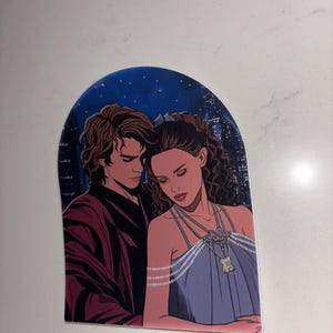 May include: A sticker featuring a romantic illustration of two figures. The man wears a burgundy jacket, and the woman is in a blue dress with a necklace. The background shows a night sky with stars and city lights.