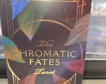 Puede incluir: La caja del mazo de tarot Chromatic Fates presenta un fondo burdeos oscuro con detalles dorados y un diseño de hojas superpuestas en tonos azules, verdes y naranjas. El título "The Chromatic Fates Tarot" está escrito en letras doradas.