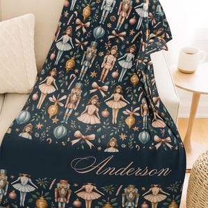 May include: A navy blue throw blanket featuring a repeating pattern of ballerinas, nutcrackers, bows, and ornaments. The blanket has the name "Anderson" printed in a cursive font at the bottom. The blanket is draped over a beige sofa.