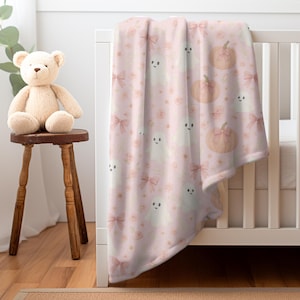 May include: A pink baby blanket with a pattern of smiling ghosts, pumpkins with bows, and pink bows and roses. The blanket is draped over a white crib, with a teddy bear on a wooden stool in the background.