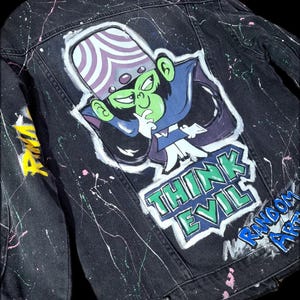Peut inclure: Veste en jean noire avec un personnage de dessin animé peint à la main au dos. Le personnage est vert avec un chapeau violet et blanc. Les mots "THINK EVIL" sont peints en vert et bleu. Détails de peinture éclaboussée en rose et blanc.
