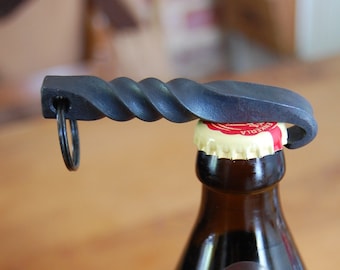 May include: A black metal bottle opener with a twisted handle and a ring on the end. The opener is being used to open a brown glass bottle of Bamberg's Spezialalt beer.