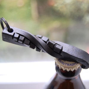 Op de afbeelding: Een zwarte metalen flesopener met een gedraaid ontwerp en een ring om op te hangen. De opener staat op een bruine glazen fles, waarbij de dop gedeeltelijk is verwijderd. De achtergrond is wazig.