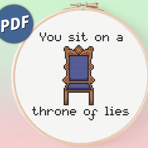 May include: A cross stitch pattern in a wooden embroidery hoop featuring a pixelated blue and brown chair centered between the black text You sit on a throne of lies. The design is displayed on a white fabric background with a blue PDF icon.