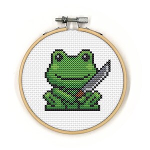 May include: A green frog holding a silver knife in its right hand. The frog is embroidered on a white canvas and framed in a wooden hoop.
