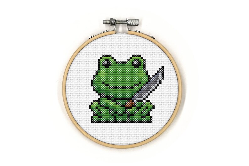 May include: A green frog holding a silver knife in its right hand. The frog is embroidered on a white canvas and framed in a wooden hoop.
