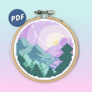 May include: A circular wooden embroidery hoop containing a cross-stitch pattern of a mountain landscape with a purple sky, a yellow sun, and dark green pine trees. A blue circular icon with the white text PDF is positioned in the top left corner.