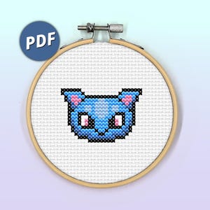 May include: A cross-stitch pattern of a blue cat face with pink ears and white eyes. The pattern is displayed in a wooden embroidery hoop.