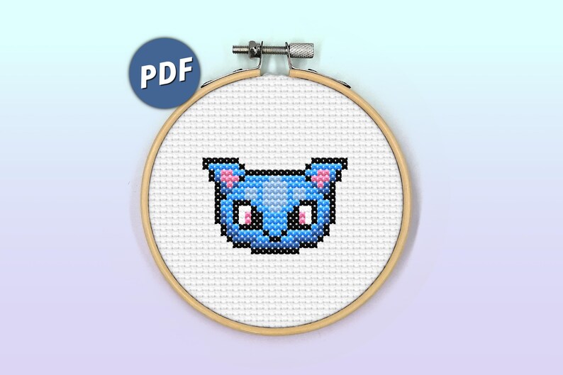May include: A cross-stitch pattern of a blue cat face with pink ears and white eyes. The pattern is displayed in a wooden embroidery hoop.