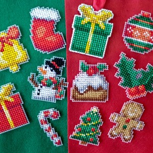 May include: A collection of colorful, pixelated Christmas ornaments. The ornaments include a bell, stocking, gift, snowman, cake, holly, candy cane, Christmas tree, gingerbread man, and a striped ball. The ornaments are on a green and red background.