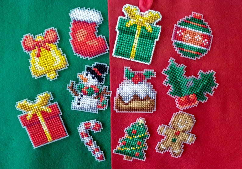 May include: A collection of colorful, pixelated Christmas ornaments. The ornaments include a bell, stocking, gift, snowman, cake, holly, candy cane, Christmas tree, gingerbread man, and a striped ball. The ornaments are on a green and red background.