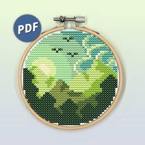 以下が含まれることがあります： 木製の円形刺繍枠に、丘のある緑の風景と、3羽の小さな黒い鳥のシルエットが描かれた明るい緑の空のクロスステッチパターンが入っています。白い文字でPDFと書かれた青い円形のアイコンが表示されています。