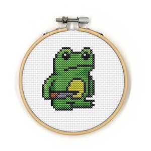 May include: A green frog with black eyes and a yellow belly is embroidered on a white fabric stretched over a wooden embroidery hoop.