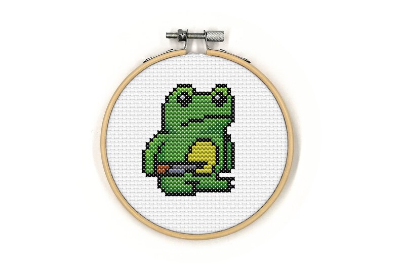 May include: A green frog with black eyes and a yellow belly is embroidered on a white fabric stretched over a wooden embroidery hoop.