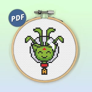 May include: A cross-stitch pattern of a green alien with four arms and a smiling face, inside a white circle. The alien is wearing a red and white hat. The pattern is in a wooden embroidery hoop.