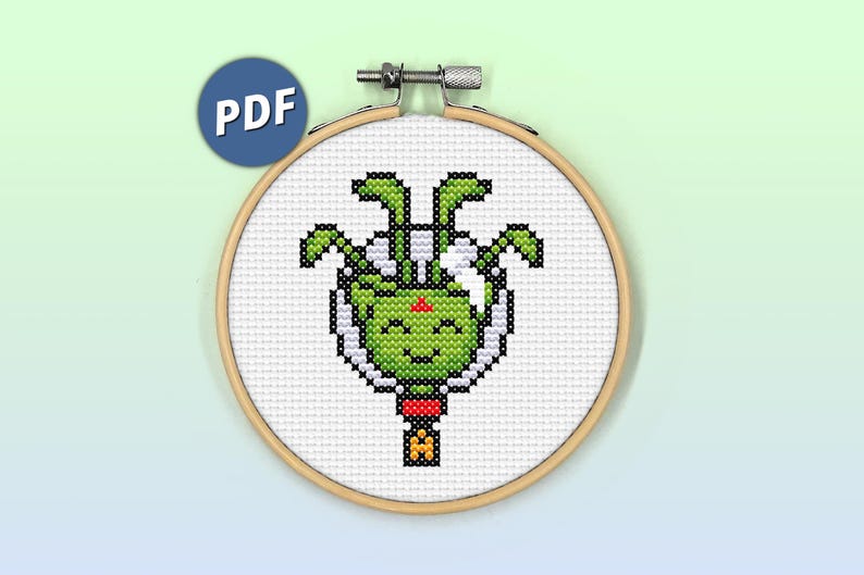 May include: A cross-stitch pattern of a green alien with four arms and a smiling face, inside a white circle. The alien is wearing a red and white hat. The pattern is in a wooden embroidery hoop.