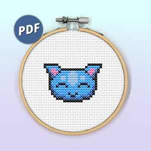 May include: A cross-stitch pattern of a blue cat with pink ears and a white face. The cat is stitched on white Aida cloth and framed in a wooden hoop.