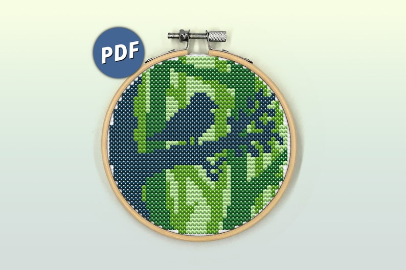 Op de afbeelding: Een ronde houten borduurring met een kruissteekproject met een gepixeld camouflagepatroon in tinten donker marineblauw, limoengroen en wit. Een blauw rond icoon met de witte tekst PDF bevindt zich in de linkerbovenhoek van de afbeelding.