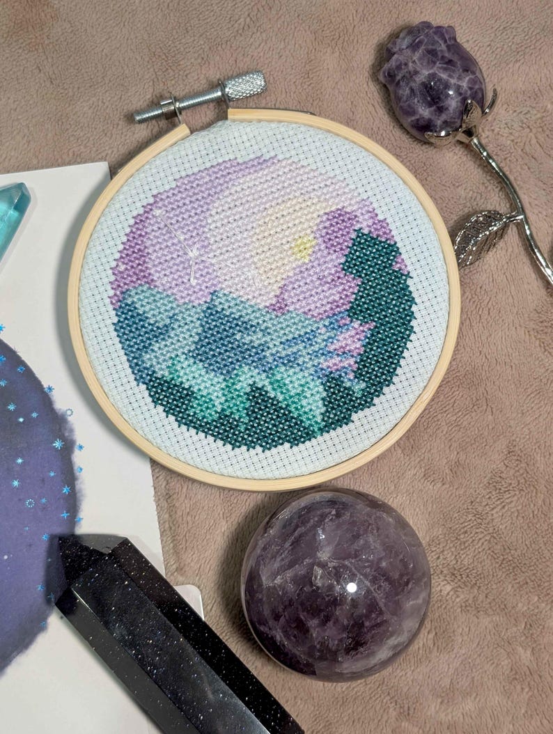May include: A circular wooden embroidery hoop containing a cross-stitch design of a landscape with a pale yellow sun, purple sky, and dark teal mountains. The fabric is white Aida cloth, and the hoop features a metal tightening screw at the top.
