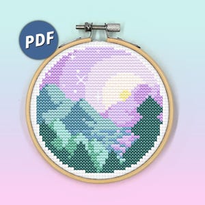 May include: A circular wooden embroidery hoop containing a cross-stitch pattern of a mountain landscape at sunset. The design features dark green pine trees, blue mountains, and a purple sky with a yellow sun and white stars. A blue PDF icon is visible.
