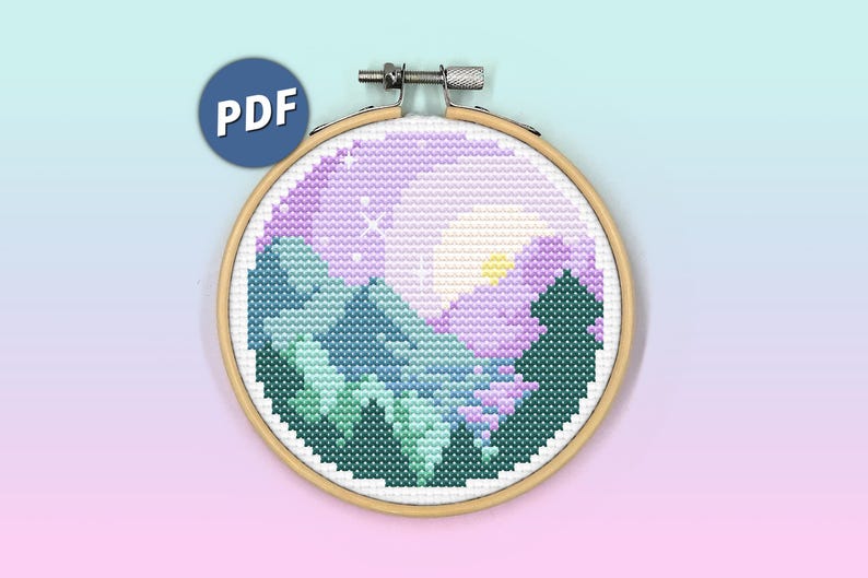 May include: A circular wooden embroidery hoop containing a cross-stitch pattern of a mountain landscape at sunset. The design features dark green pine trees, blue mountains, and a purple sky with a yellow sun and white stars. A blue PDF icon is visible.