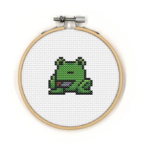 May include: A green frog with a brown eye and a brown mouth is embroidered on a white fabric stretched over a wooden embroidery hoop.