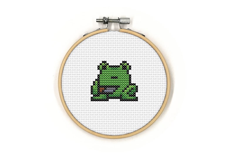 May include: A green frog with a brown eye and a brown mouth is embroidered on a white fabric stretched over a wooden embroidery hoop.