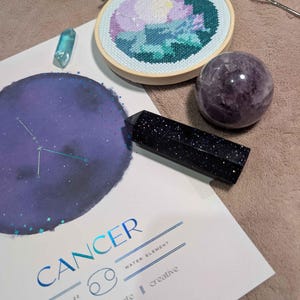 May include: A Cancer zodiac print featuring a purple moon and constellation, a round wooden embroidery hoop with a floral cross-stitch, a dark blue goldstone crystal tower, a purple sphere, a small light blue crystal, and a metal rose ornament.