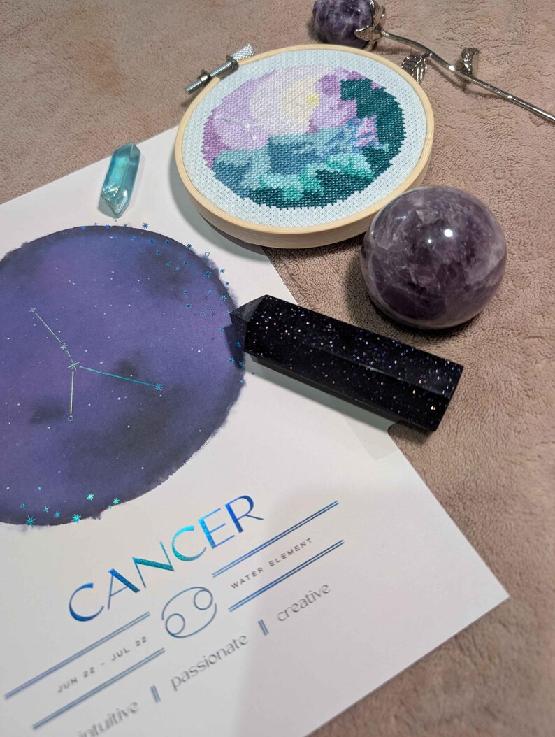 May include: A Cancer zodiac print featuring a purple moon and constellation, a round wooden embroidery hoop with a floral cross-stitch, a dark blue goldstone crystal tower, a purple sphere, a small light blue crystal, and a metal rose ornament.