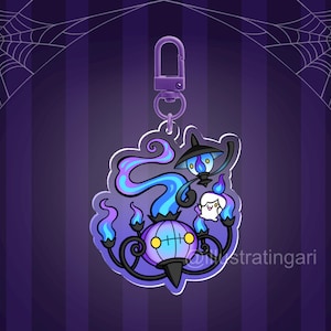 Puede incluir: Un charm de acrílico morado y azul con un diseño de fantasma y lámpara de dibujos animados. El charm tiene un cierre morado y está sobre un fondo a rayas moradas con una telaraña.