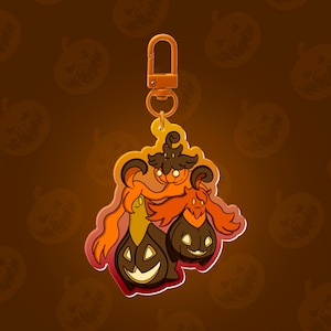 May include: A Halloween-themed keychain featuring a pumpkin-headed character with a witch hat and two pumpkin companions. The design incorporates orange, black, and yellow colors, with a gold-colored clasp. The background is a gradient of brown with pumpkin silhouettes.