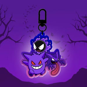 Puede incluir: Un llavero morado con tres personajes de fantasmas de dibujos animados con ojos rojos y una larga lengua rosa. El fondo es un degradado de púrpura con siluetas de árboles y murciélagos.