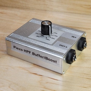 May include: Silver audio effects pedal with a black knob and frequency markings. The text "Piezo HPF Buffer/Boost" is visible on the front. Includes input and output jacks. Measures approximately 10 cm x 7 cm.