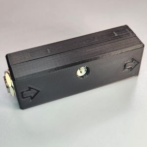 May include: A black rectangular device with a small circular opening and two arrow symbols. The device has a metallic component on one end. The object is likely a component or part of a larger system.