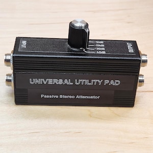 May include: A black rectangular Universal Utility Pad passive stereo attenuator with a silver and black rotary knob on top. The device features input and output labels, a decibel scale from -10dB to -40dB, and four metal ports on the sides.