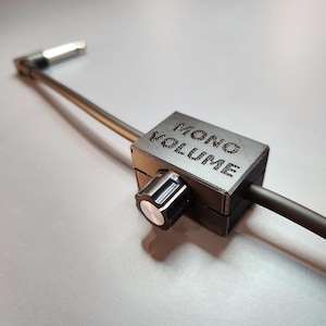 Könnte beinhalten: Ein schwarzer Kunststoff-Mono-Lautstärkeregler mit einem silbernen Knopf und einem daran befestigten schwarzen Kabel. Der Knopf ist mit "MONO VOLUME" beschriftet.