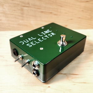 Peut inclure: Une pédale de sélection de ligne double verte avec un interrupteur au pied argenté et deux prises d'entrée. La pédale porte l'inscription "DUAL LINE SELECTOR" sur le dessus.