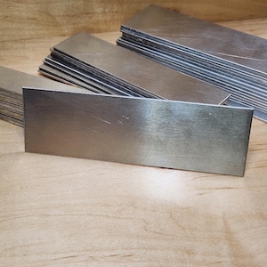 May include: Stacks of rectangular, brushed metal plates. The plates are silver-colored and appear to be made of a solid metal. The plates are arranged in neat stacks on a light brown wooden surface.