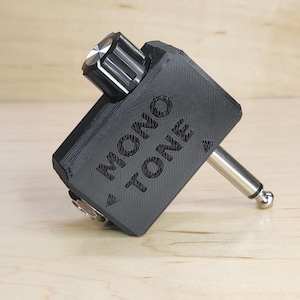 May include: A black 3D-printed guitar accessory featuring a silver-topped black control knob and a metal 1/4-inch jack plug. The rectangular housing is embossed with the text MONO TONE and includes small triangular directional indicators.