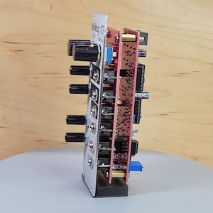 May include: A modular synthesizer module with a silver faceplate, black knobs, and a red circuit board. The module has several knobs and switches, and the text "MinMod VCF" is visible. The module is designed for electronic music production.
