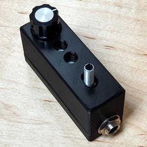 May include: A black electronic device with a silver knob, three holes, a silver cylinder, and a silver jack. The device is rectangular and appears to be 3D-printed. The knob has a silver top and a black base.