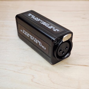 Puede incluir: Un adaptador de audio negro con conector XLR. El dispositivo tiene texto que dice "BALANCED XLR PAD -20dB" y "PHANTOM: PASS (P48 OK)". El adaptador es rectangular y tiene una pieza metálica plateada en el lateral.
