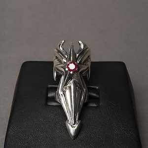 May include: A silver ring with a star design and a red gemstone in the center. The ring is on a black display stand.