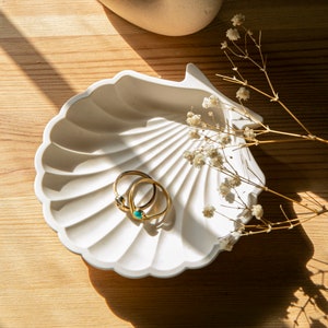 May include: A white, shell-shaped trinket dish holds two gold rings, one with a teal stone and the other with a dark stone. Dried white flowers and a light wooden surface complete the scene.