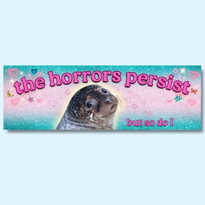 Könnte beinhalten: Rechteckiges Banner mit hellblauem Hintergrund und einem glitzernden Türkis- und Rosa-Farbverlauf. Das Banner zeigt eine Robbe mit dem Text "the horrors persist" und "but so do I" in Rosa.