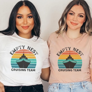 May include: Two women wearing t-shirts with a retro sunset graphic and the text "Empty Nest Cruising Team". One shirt is white and the other is pink.