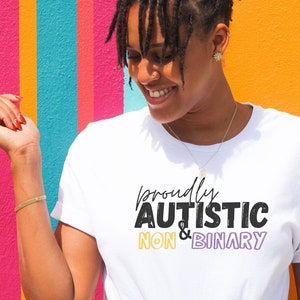 May include: A white t-shirt with the text "proudly autistic non-binary" in black, yellow, and purple lettering. The person wearing the shirt is smiling and standing in front of a colorful striped wall.