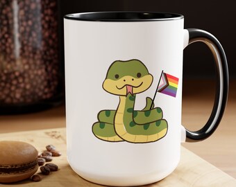 May include: White ceramic mug with a black rim and handle. The mug features a cartoon green snake holding a rainbow pride flag.