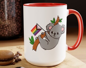 May include: White ceramic mug with a red rim and handle. The mug features a cartoon koala holding a rainbow pride flag while hanging from a tree branch.